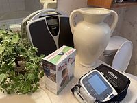 Photo showing Salter scale, Magic Bag box, Omron blood pressure monitor with cuff and case, artificial plant, and white ceramic vase in the background.