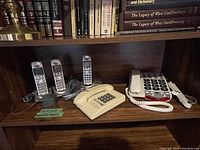 Telephones displayed on a wooden shelf showing three cordless handsets on a charging base, a beige wired phone, and a large-button phone.