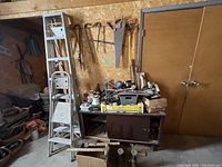 Wide image showing metal step ladder, multiple hand saws hanging on wooden wall, cabinet with cluttered tools and hardware supplies on top.