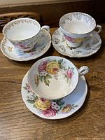Set of three vintage porcelain cups and saucers with floral and gold leaf designs, viewed from the side showing outer decoration.