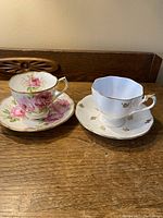Two porcelain cups and saucers on wood surface, one set with pink roses, the other white with gold leaf motif.