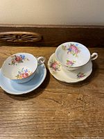 Two vintage teacups and their matching saucers on a wooden surface, showing floral designs and gold trim.
