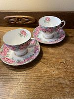 Photo showing two cups with their matching saucers, placed on a wooden surface with visible carved wood detail. Both cup and saucer have pink rose floral patterns with gold trim.
