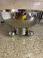 Front view showing the polished stainless steel bowl with three ball-shaped legs and ribbed base, placed on a speckled countertop with tiled backsplash.