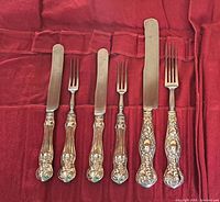 Full shot of all 6 antique sterling silver cutlery pieces displayed on red fabric, showing varying sizes and ornate handles