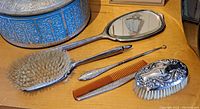 Vanity set pieces arranged on a wooden surface, showing the hand mirror, hair brush, button hook, comb, and clothes brush with silver detailing.