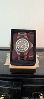 Frontal angle photo of the wooden watch on a cushion inside black base stand with Tense label