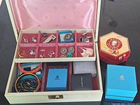 Open vintage jewelry box showing pink velvet compartments holding earrings, rings, brooch, and large silver tone earrings, also showing some boxes including Birks and George Jensen and a decorative Austrian Mozarte Kugeln box with bead jewelry inside.