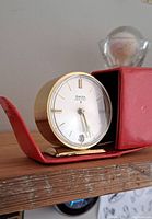 Vintage Swiza travel clock with white dial, gold-tone markers and hands, and date window, positioned inside red leather carry case on wooden surface.