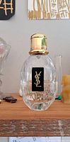 Clear faceted glass YSL fragrance bottle with gold cap on a shelf, showing front label with YSL logo.