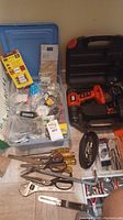Photo showing the drill carrying case, assorted hardware like screws, hooks, and various hand tools including scissors and pliers laid out on floor.