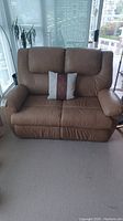 Front view of tan leather 2-seater recliner loveseat with decorative pillows on seat cushions, situated near large windows.