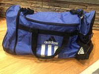 Full view of blue Adidas duffel bag on wood floor showing logo, shape, side pockets and straps