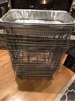 Stack of 25 large rectangular aluminum wire-frame chafing dishes with aluminum pans inside, stacked on a hardwood floor with a stone fireplace background.