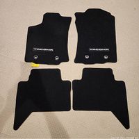 Four black carpeted floor mats arranged on light carpet, two front mats have 'TACOMA' logo and metal grommets visible; driver mat shows light wear.