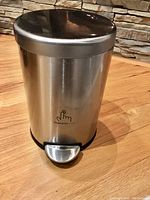 Round stainless steel pedal trashcan shown from front with visible Simple Human logo and foot pedal on wooden floor background.