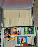 Photo of a large open box filled with stacks of loose sports cards of various sizes and thicknesses, with visible unopened packs and a 1990 BOX H2 Series 1 pack.