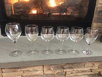 Front row of six assorted clear wine glasses, five about 7 inches tall, one about 8 inches tall, arranged on stone hearth with fireplace background.