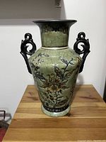 Green textured ceramic vase with floral and bird decoration, two ornamental handles, displayed on wooden table.