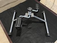 Silver metal bicycle peddle style exercise stand with two black plastic pedals and rubber feet shown on black surface