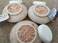 Three white plastic containers with coarse pink Himalayan bath salt crystals visible inside and lids on the side, each tagged with a printed label card.