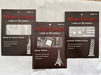 Photo of the three Model Technics laser cut 3D architectural model packages: Eiffel Tower, Bank of China Tower, and Leaning Tower of Pisa. Shows front of packaging with age rating 14+. Provides brand and product details.