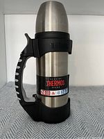 Front and side view of a stainless steel Thermos bottle with black handle and base, showing the label indicating 24 hours hot and cold retention.
