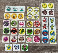 Flat lay photo showing multiple vintage scratch and sniff stickers arranged in rows, featuring Watson's ointment, ice cream cones, Halloween ghosts, dinosaur, monkey faces, cartoon characters, crabs, and fruits.