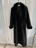 Front view of vintage black women's full-length winter coat with faux fur collar and cuffs, hanging on a door.