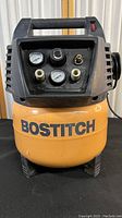 Front view of Bostitch air compressor showing control panel, two pressure gauges, regulator knob, and air hose connectors. Yellow tank labeled with brand.