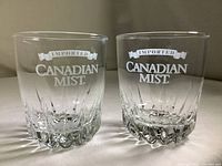 Two clear collectible Canadian Mist whiskey glasses side by side on a white surface, showing front branding and pattern detail.