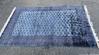 Full view of blue and purple geometric pattern area rug on asphalt surface, showing the complete design and fringe on ends.