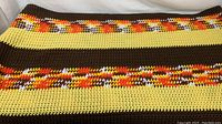 Handmade crochet blanket laid flat showing horizontal stripes in brown, yellow, and multicolored yarns.
