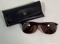 Black leather case with button snap closure and Maui Jim tortoise shell sunglasses with brown lenses laid out flat.