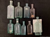 Overview of nine vintage glass medicine bottles in multiple shapes and colors on black background