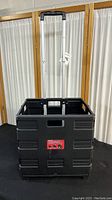Front view of the black foldable rolling crate with extended handle and label on front.