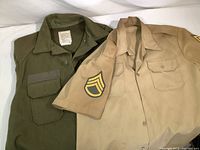 Photo of folded olive green US Army winter jacket next to Staff Sergeant khaki shirt showing the rank insignia patch on the shirt's sleeve.