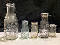 Five vintage food and cream glass bottles arranged in ascending size order on black fabric background. Clear to lightly tinted glass with embossed writing and logos visible.