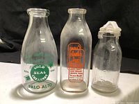 Three vintage glass milk bottles lined up against a black background. From left to right: Gold Seal Creamery Palo Alto quart bottle with green painted label, Carmel Dairy quart bottle with orange printed label, Absolutely Pure Milk embossed bottle with glass top taped.