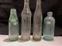 Four vintage soda bottles standing side by side against a dark background, showing differing heights and designs including a marble stopper and varying embossments.