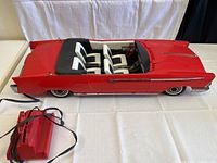 Front angled view of vintage red mechanical toy car with four white and black seats and wired remote control beside it.