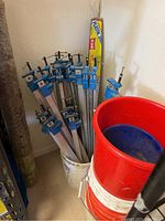 Bucket containing multiple aluminum pipe clamps with blue clamp heads and handles, with red bucket in foreground and plastic sheeting roll behind