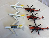 Overview photo showing three white passenger planes with yellow tails and three red helicopters with black and red blades, arranged on white surface.