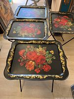 Four black vintage metal folding TV trays with colorful floral designs and gold leaf borders worked out and displayed on a floor.