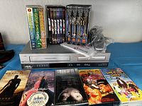 Philips VHS DVD Player combo with remote, cables, and various VHS tapes and DVDs displayed in front and on top.