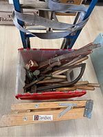 Top view of red plastic bin filled with various vintage saws and hand tools placed in front of refrigerator dolly and Davidson wooden step ladder