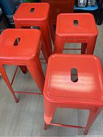 Top angle view showing four red-orange metal stools with square tops and cutout handles.