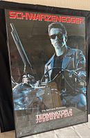 Framed Terminator 2 Judgment Day movie poster leaning against a wall, clearly showing Arnold Schwarzenegger holding a shotgun on a motorcycle with a black background and red text.