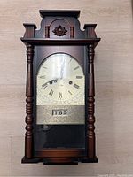 Full frontal view of the vintage wooden wall clock showing columns, ornate top, metal face with Roman numerals, clock hands, and calendar window.