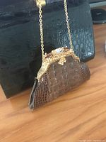 Brown evening lipstick bag with crocodile texture and decorative gold-tone frame and chain strap resting beside the black handbag.